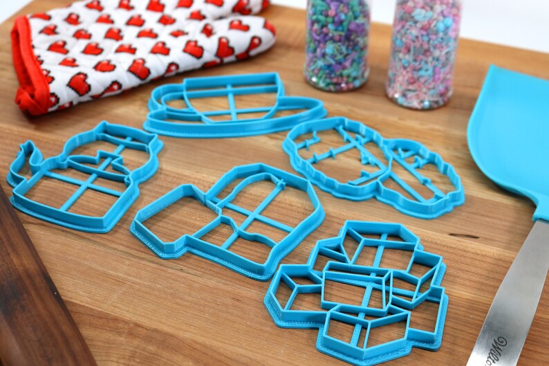 May include: A set of six blue plastic cookie cutters shaped like a teapot, mug, cup and saucer, a snowflake, a house, and a gift box. The cutters are on a wooden surface.