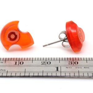 May include: A pair of orange stud earrings with a unique design. The earrings are approximately 1 cm in diameter and are shown next to a ruler.