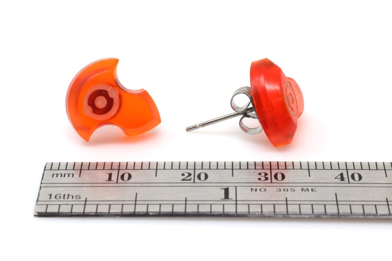 May include: A pair of orange stud earrings with a unique design. The earrings are approximately 1 cm in diameter and are shown next to a ruler.