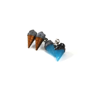 May include: A pair of earrings featuring miniature ice cream cones and a blue, heart-shaped design. The ice cream cones have brown cones and gray tops. The heart-shaped earring is blue with black accents. The earrings are on a white background.