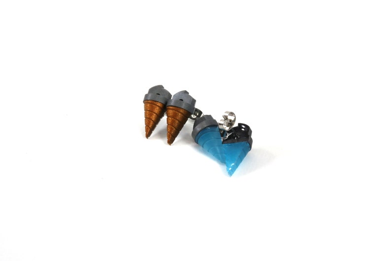 May include: A pair of earrings featuring miniature ice cream cones and a blue, heart-shaped design. The ice cream cones have brown cones and gray tops. The heart-shaped earring is blue with black accents. The earrings are on a white background.