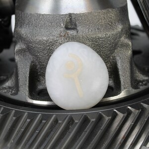 May include: A white stone with a symbol etched into it, resting on a metal gear. The symbol is a circle with a crescent moon and a staff.