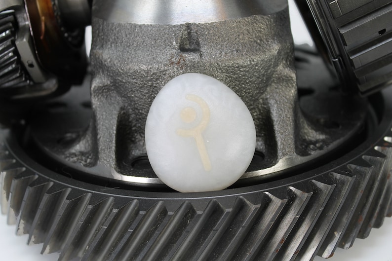 May include: A white stone with a symbol etched into it, resting on a metal gear. The symbol is a circle with a crescent moon and a staff.