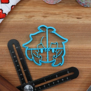 May include: A blue plastic cookie cutter shaped like a cartoon character with a large head and short hair. The character has large eyes and a small mouth.