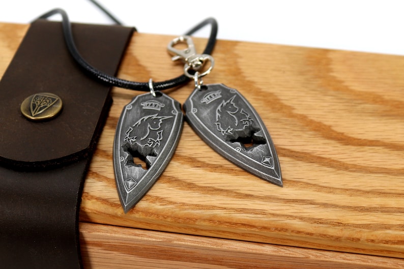 May include: Two silver pendants in the shape of a shield with a unicorn design. The pendants are on a wooden surface with a leather pouch in the background.