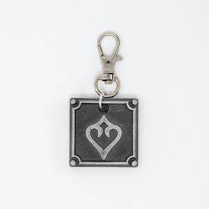 May include: A silver keychain with a square charm featuring a stylized heart design. The charm has a silver border and a black background.