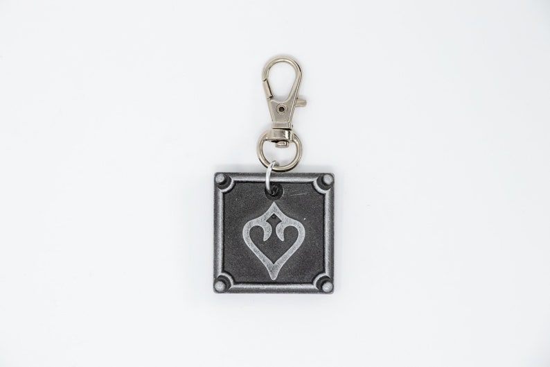 May include: A silver keychain with a square charm featuring a stylized heart design. The charm has a silver border and a black background.