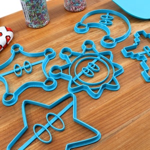 May include: A set of six blue plastic cookie cutters in various shapes, including a star, a sun, a crown, a spaceship, and a Christmas tree. The cutters are on a wooden surface.