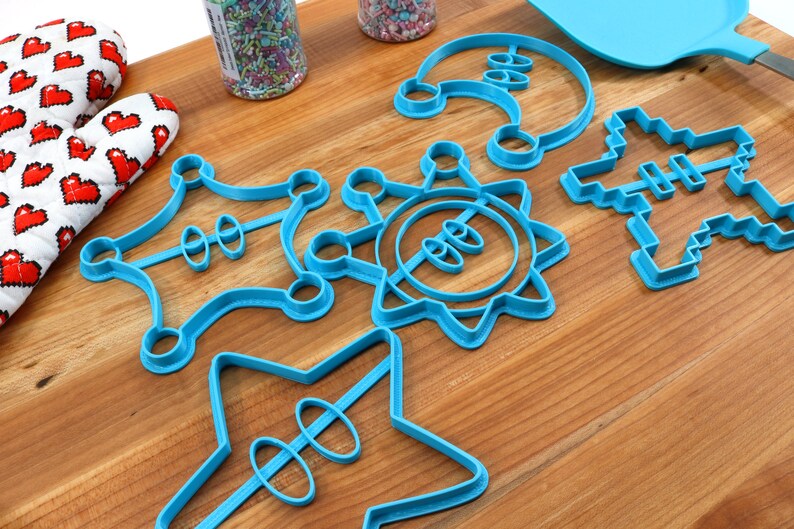 May include: A set of six blue plastic cookie cutters in various shapes, including a star, a sun, a crown, a spaceship, and a Christmas tree. The cutters are on a wooden surface.