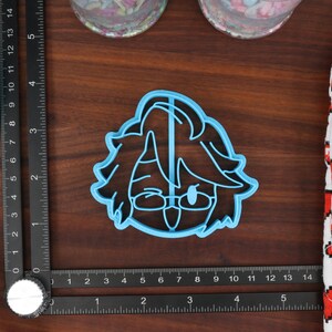 May include: A blue plastic cookie cutter shaped like a cartoon character with brown hair, glasses, and a tongue sticking out.