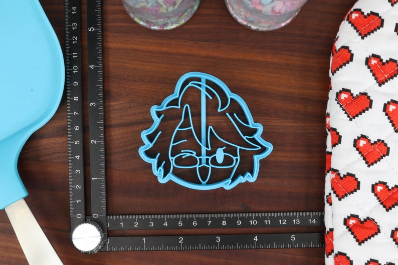 May include: A blue plastic cookie cutter shaped like a cartoon character with brown hair, glasses, and a tongue sticking out.
