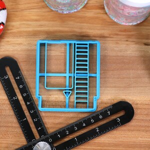May include: A blue plastic cookie cutter shaped like a vintage video game cartridge. The cutter has a detailed design with a slot, a label, and a ladder on the side.