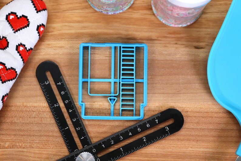 May include: A blue plastic cookie cutter shaped like a vintage video game cartridge. The cutter has a detailed design with a slot, a label, and a ladder on the side.