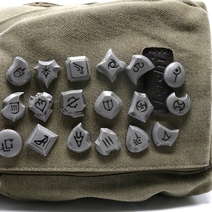 May include: A close-up of a gray canvas bag with 18 silver metal pins featuring various symbols. The pins are arranged in a grid pattern on the bag.