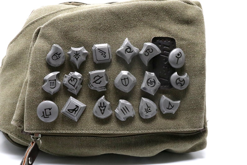 May include: A close-up of a gray canvas bag with 18 silver metal pins featuring various symbols. The pins are arranged in a grid pattern on the bag.