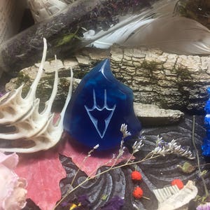 May include: A blue glass pendant with a white stylized design of a three-pronged symbol. The pendant is surrounded by dried flowers and other natural elements.