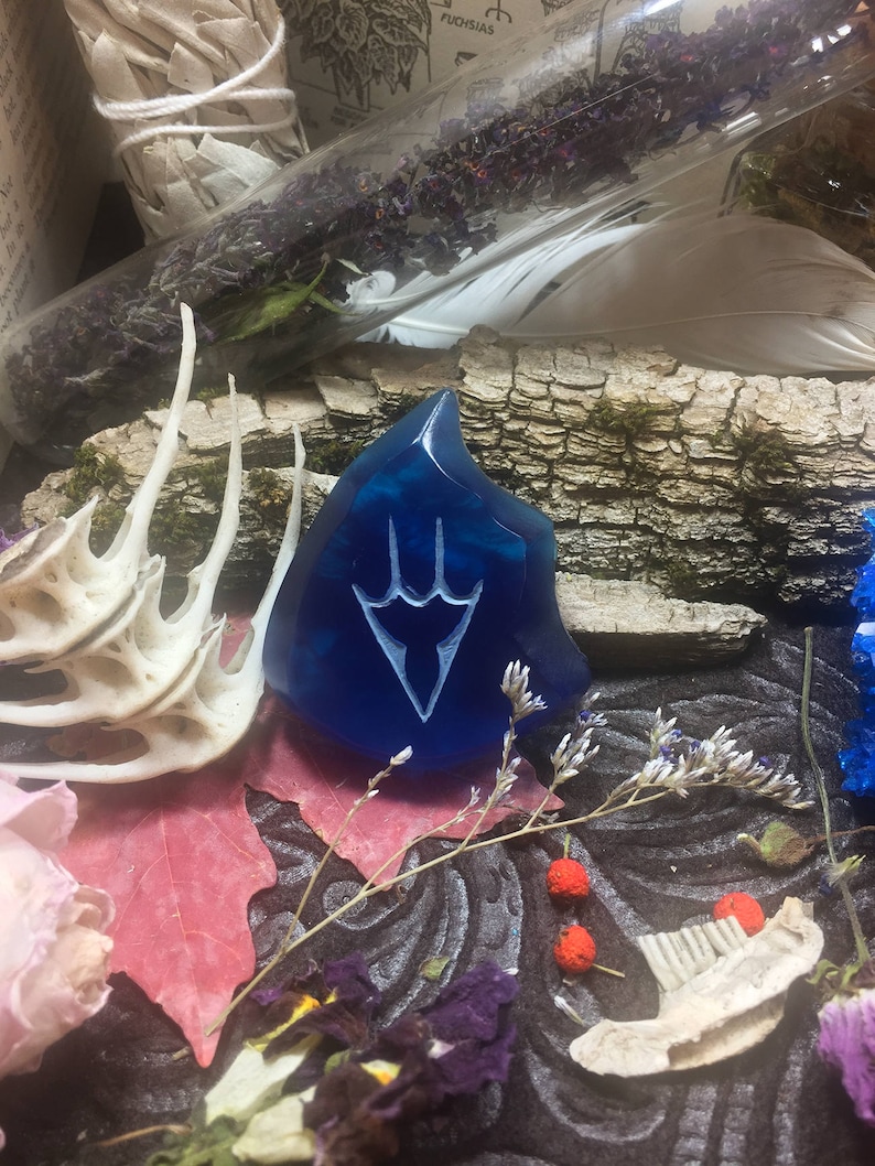 May include: A blue glass pendant with a white stylized design of a three-pronged symbol. The pendant is surrounded by dried flowers and other natural elements.