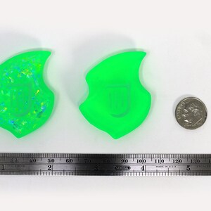 Op de afbeelding: Twee groene harsvormen, een met een glinsterende afwerking en de andere met een gladde afwerking. De vormen zijn vlamvormig en ongeveer 5 cm hoog. Een Amerikaanse dime is opgenomen ter vergelijking.