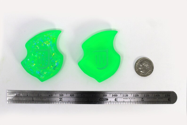 Op de afbeelding: Twee groene harsvormen, een met een glinsterende afwerking en de andere met een gladde afwerking. De vormen zijn vlamvormig en ongeveer 5 cm hoog. Een Amerikaanse dime is opgenomen ter vergelijking.