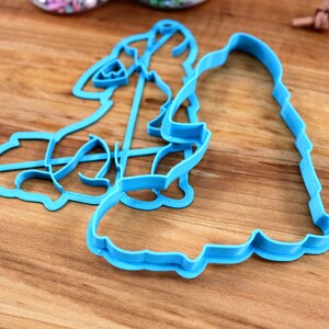 May include: Two blue plastic cookie cutters shaped like a woman and a man. The cutters are on a wooden surface.