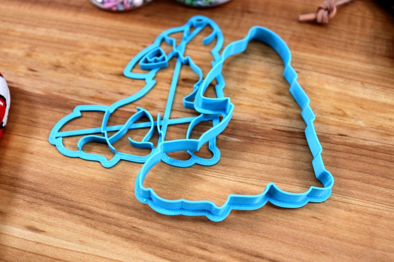 May include: Two blue plastic cookie cutters shaped like a woman and a man. The cutters are on a wooden surface.
