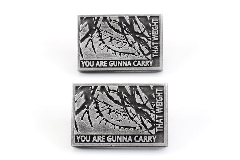 May include: Two silver metal pins with a distressed, textured design. The pins feature the text "You are gunna carry that weight" in black.