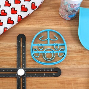 May include: A blue plastic cookie cutter shaped like a cartoon face with closed eyes, a nose, and a mouth. The cookie cutter is on a wooden surface.