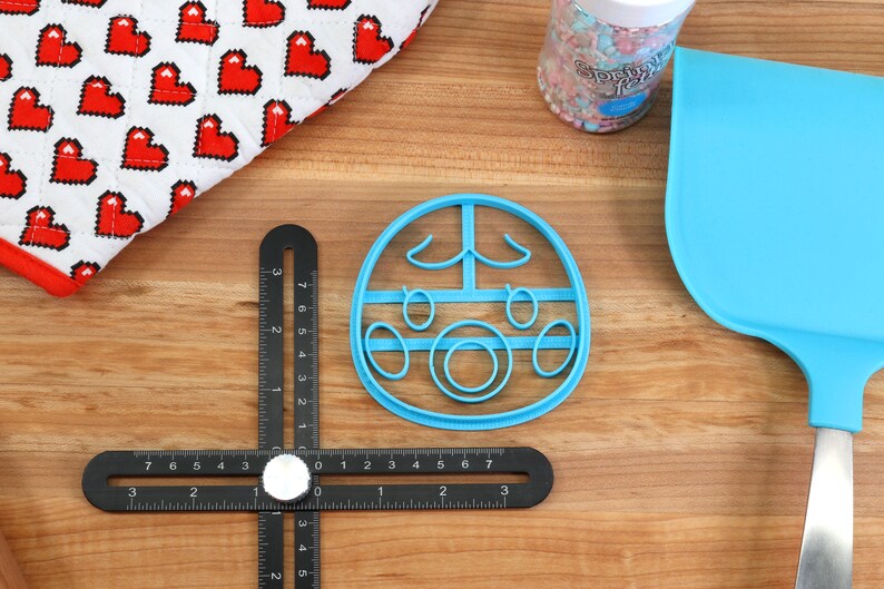 May include: A blue plastic cookie cutter shaped like a cartoon face with closed eyes, a nose, and a mouth. The cookie cutter is on a wooden surface.