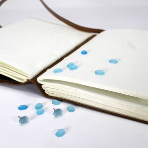 K&ouml;nnte beinhalten: Ein braunes, ledergebundenes Tagebuch mit blauen Glasakzenten, die auf den Seiten und um den Einband verstreut sind. Das Tagebuch ist ge&ouml;ffnet und die Seiten sind leer.