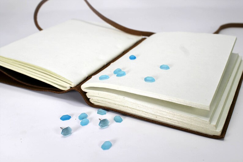 K&ouml;nnte beinhalten: Ein braunes, ledergebundenes Tagebuch mit blauen Glasakzenten, die auf den Seiten und um den Einband verstreut sind. Das Tagebuch ist ge&ouml;ffnet und die Seiten sind leer.