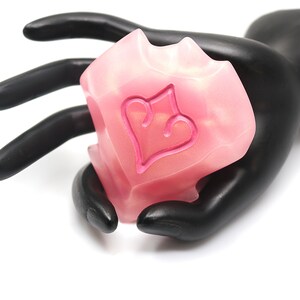 May include: A pink, heart-shaped decorative object with a heart design, held by a black hand. The object has a translucent appearance and a textured surface. The heart design is a darker shade of pink.