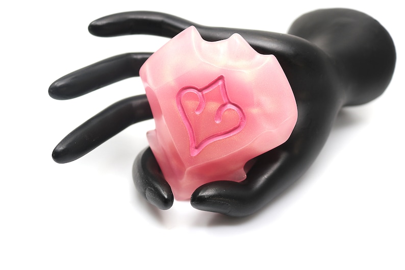 May include: A pink, heart-shaped decorative object with a heart design, held by a black hand. The object has a translucent appearance and a textured surface. The heart design is a darker shade of pink.