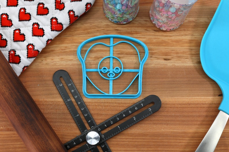 May include: A blue plastic cookie cutter shaped like a slice of bread with a cartoon face. The cutter has a grid pattern and a small nose and eyes.