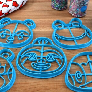 May include: Five blue plastic cookie cutters shaped like animal faces. The cutters are on a wooden surface. The cutters are for making cookies in the shapes of a panda, a frog, a sloth, a bear, and a turtle.