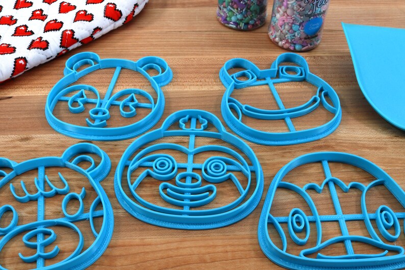 May include: Five blue plastic cookie cutters shaped like animal faces. The cutters are on a wooden surface. The cutters are for making cookies in the shapes of a panda, a frog, a sloth, a bear, and a turtle.