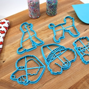 May include: A set of six blue plastic poodle cookie cutters on a wooden surface. The cutters are in various shapes and sizes, including a poodle with a hat, a poodle with a bow, and a poodle with a tail.