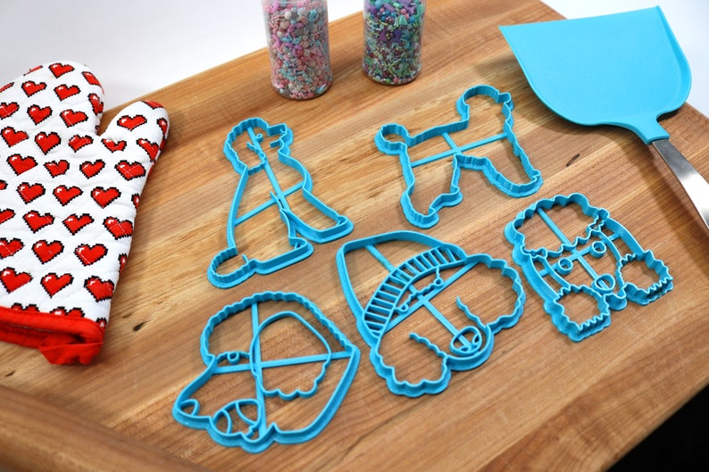 May include: A set of six blue plastic poodle cookie cutters on a wooden surface. The cutters are in various shapes and sizes, including a poodle with a hat, a poodle with a bow, and a poodle with a tail.