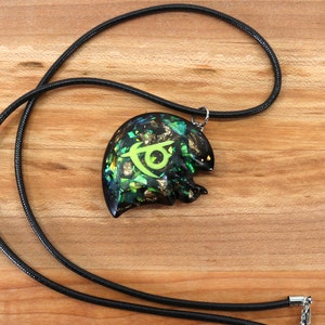 May include: A black cord necklace with a green and gold pendant featuring a stylized symbol. The pendant is made of resin and has a glittery finish.