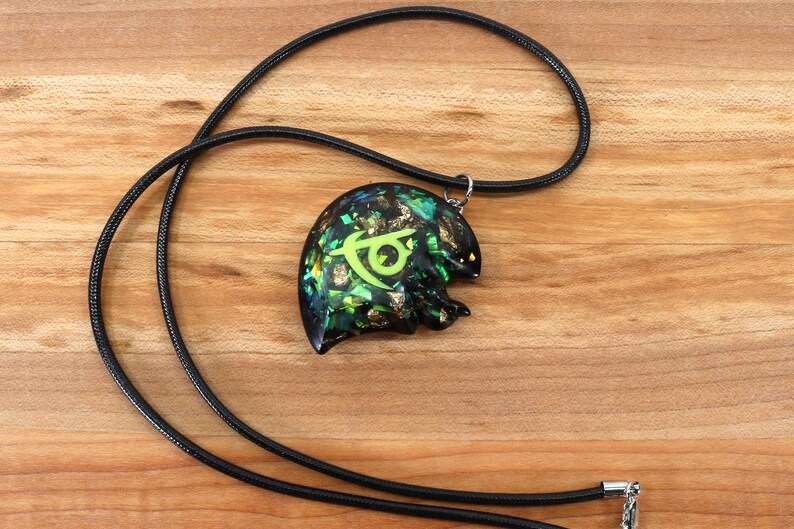 May include: A black cord necklace with a green and gold pendant featuring a stylized symbol. The pendant is made of resin and has a glittery finish.