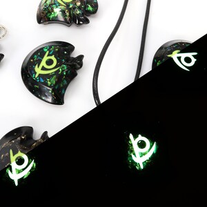 May include: A set of four black resin pendants with a green and gold glitter design. The pendants feature a stylized eye symbol. The pendants are shown in both regular and glow-in-the-dark lighting.