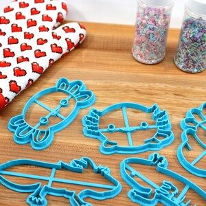 May include: Five blue plastic cookie cutters shaped like axolotls, a crab, and a hedgehog. The cutters are on a wooden surface with two jars of sprinkles in the background.