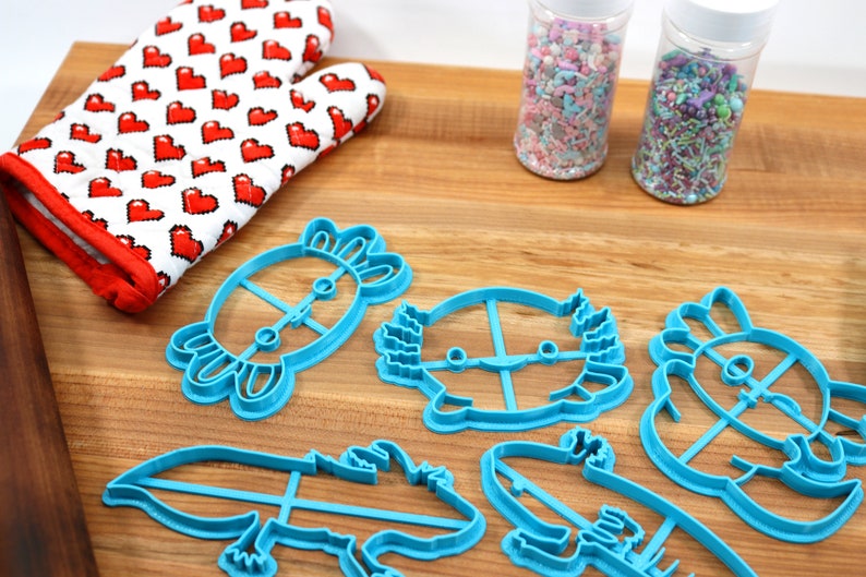 May include: Five blue plastic cookie cutters shaped like axolotls, a crab, and a hedgehog. The cutters are on a wooden surface with two jars of sprinkles in the background.