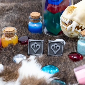 May include: Two silver metal square cufflinks with a decorative design of a stylized heart. The cufflinks are on a brown furry surface with small glass bottles filled with colored liquids and a small animal skull.