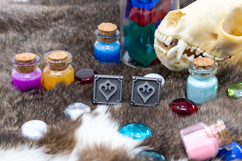 May include: Two silver metal square cufflinks with a decorative design of a stylized heart. The cufflinks are on a brown furry surface with small glass bottles filled with colored liquids and a small animal skull.