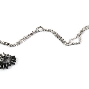 May include: A silver chain necklace with a black metal pendant featuring a stylized mask with sharp edges and two large eyes.