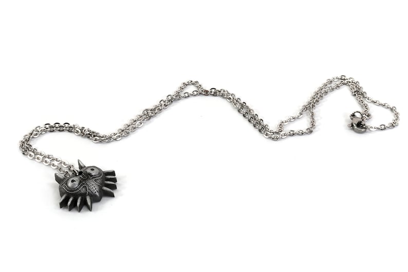 May include: A silver chain necklace with a black metal pendant featuring a stylized mask with sharp edges and two large eyes.