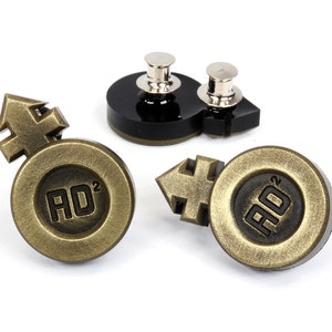 May include: Two gold-toned metal pins with a black background. Each pin features a stylized arrow symbol and the text "AD2".