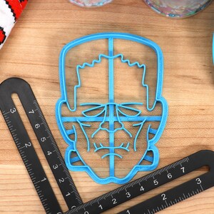 May include: A light blue plastic cookie cutter in the shape of a monster's face, with detailed features. Two black rulers are positioned around the cutter. A white cloth with red pixelated hearts is in the background.