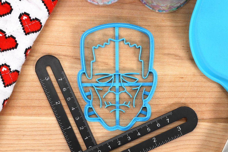 May include: A light blue plastic cookie cutter in the shape of a monster's face, with detailed features. Two black rulers are positioned around the cutter. A white cloth with red pixelated hearts is in the background.