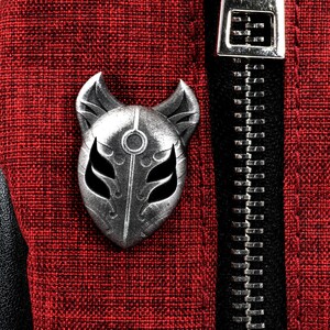 Op de afbeelding: Een zilveren metalen pin in de vorm van een vossenmasker met een zwart-wit ontwerp. De pin is bevestigd aan een rode stoffen achtergrond.
